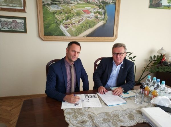 W cetnrum burmistrz Włodzimierz Badurka oraz Marcin Wojniak podpisujący dokumenty. Obaj panowie patrzą się w obiektyw aparatu. Siedzą przy stole, na którym znajdują się wody i soki. Za nimi na ścianie wiszą obrazy.