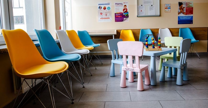 Poczekalnia w przychodni, widoczne plastikowe krzesła dla pacjentów w kolorach żółtym, białym oraz niebieskim. Obok stoi stolik dla dzieci z zabawką dookoła, którego stoją krzesełka koloru różowego, niebieskiego i zielonego.